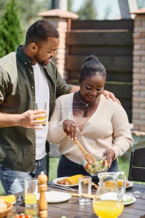happy african american man hugging wife holding salad in glass bowl while having bbq on backyard