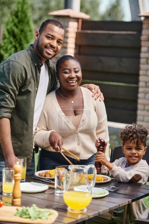 Foto de Retrato de padres afroamericanos felices e hijo preparando mesa de jardín para el almuerzo en el patio trasero - Imagen libre de derechos