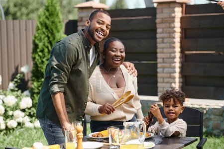 Portrait fröhlicher afrikanisch-amerikanischer Eltern und ihres Sohnes bei der Zubereitung von Gartentischen für das Mittagessen im Hinterhof