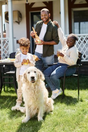 cute dog looking at camera and sitting on lawn near african american family during bbq on backyard