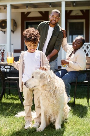 fröhlicher afrikanisch-amerikanischer Junge lächelt und streichelt Hund, während er ein Glas Orangensaft in der Nähe der Eltern hält