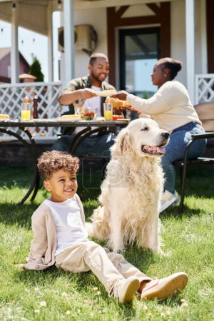 fröhlicher afrikanisch-amerikanischer Junge sitzt auf grünem Rasen neben Hund und Eltern beim Essen im Garten