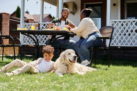 glücklich afrikanisch-amerikanisches Kind sitzt auf grünem Rasen neben Hund und Eltern beim Mittagessen im Garten