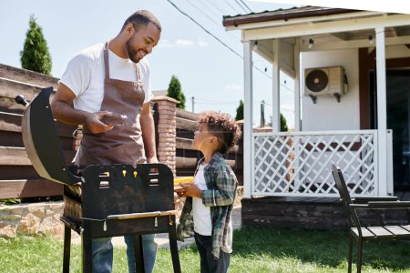 happy african american man in apron cooking corn on bbq grill and looking at son on backyard