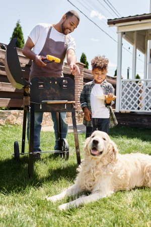 positive african american man in apron cooking corn on bbq grill and looking at dog near son