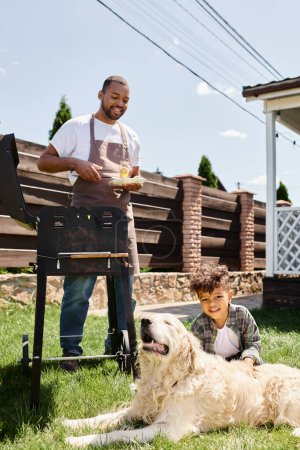 smiling african american man in apron cooking corn on bbq grill and looking at son on backyard