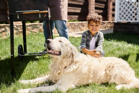 smiling african american boy sitting near dog while father cooking bbq grill on backyard in suburbs