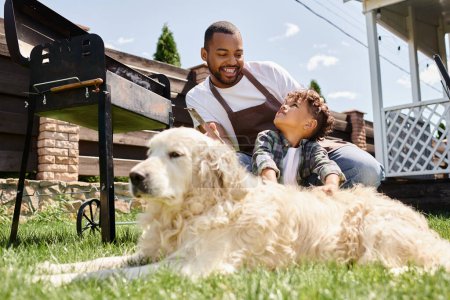 happy african american man in apron holding cooking tongs and looking at family dog near son