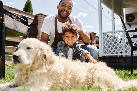 glücklich afrikanisch-amerikanischer Mann in Schürze berührt lockiges Haar seines Sohnes und sitzt neben Familienhund im Freien