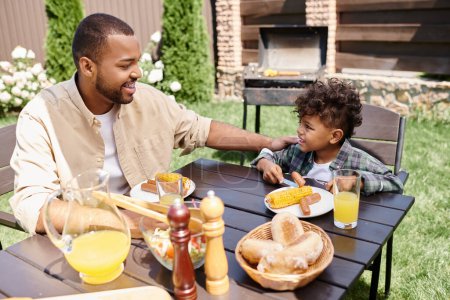 fröhlich afrikanisch-amerikanischer Vater isst Mittagessen mit lockigem Sohn im Freien, Grillwürste und Mais