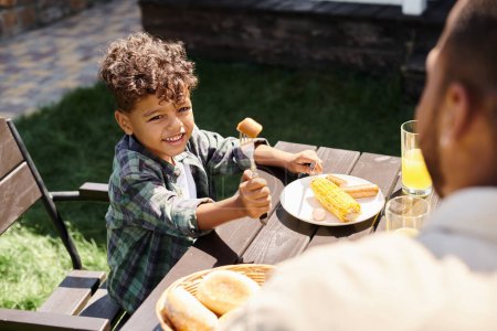 alegre rizado afroamericano chico comiendo salchichas y maíz a la parrilla mientras mira a padre al aire libre