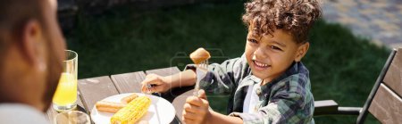 alegre rizado afroamericano chico comiendo salchichas y maíz a la parrilla mientras mira padre, pancarta