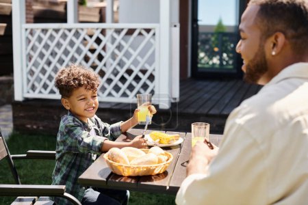 joyful african american boy eating grilled bbq meal and holding orange juice near father on backyard