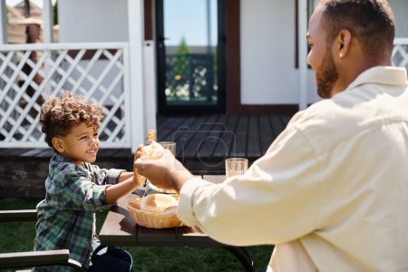 glücklich afrikanisch-amerikanischer Vater reicht frisches Brötchen an lockig lächelnden Sohn, während er Familie grillt