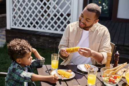 alegre afroamericano padre tener familia barbacoa con rizado hijo en patio trasero de la casa, a la parrilla de maíz