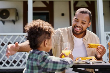 positive african american man having family bbq with curly son on backyard of house, grilled corn