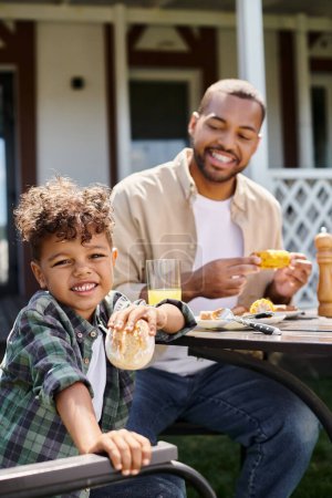 cheerful african american kid having family bbq with father on backyard of house, holding bun