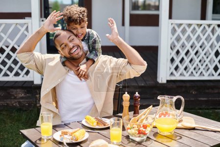 positive african american kid hugging father while having bbq lunch on backyard of house, family