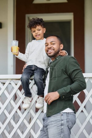 happy african american father in braces holding orange juice and hugging his son sitting on porch