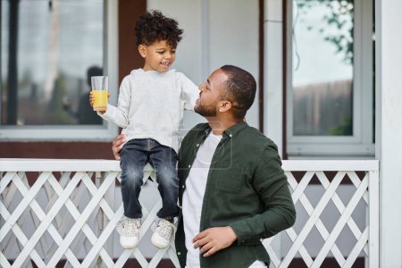 freudiger afrikanisch-amerikanischer Mann in Zahnspange, der Orangensaft hält und seinen Sohn auf der Veranda umarmt