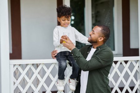 glücklich afrikanisch-amerikanischer Vater in Zahnspange hält Orangensaft und umarmt niedlichen Sohn sitzt auf der Veranda