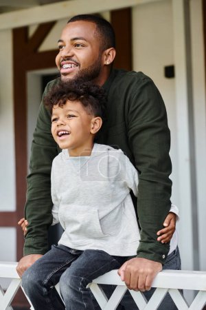 joyful african american boy sitting on porch and embracing his father in braces on backyard of house