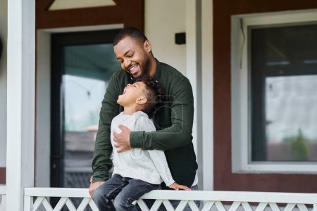fröhliche afrikanisch-amerikanische Junge sitzt auf Veranda und umarmt mit Vater in Zahnspange auf Hinterhof
