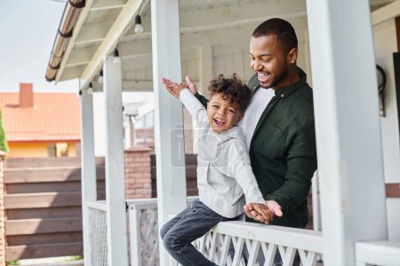 happy moments, african american boy sitting on porch and holding hands with joyful father in braces