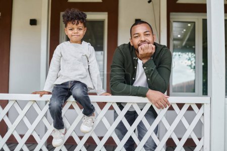 joyful african american father and son smiling and sitting on porch of house, family portrait