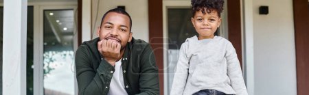 joyful african american father and son smiling and sitting on porch of house, family banner