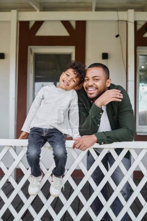 positive african american father and son smiling and sitting on porch of house, family portrait
