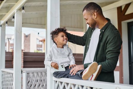 joyful african american father touching curly hair of son while sitting on porch of house, family