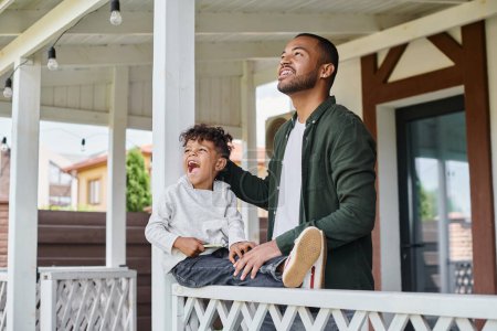 excited african american father and son smiling and sitting on porch of house, family portrait