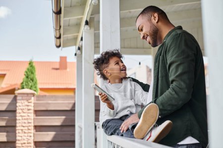 joyful african american father and son smiling and sitting on porch of house, holding smartphone