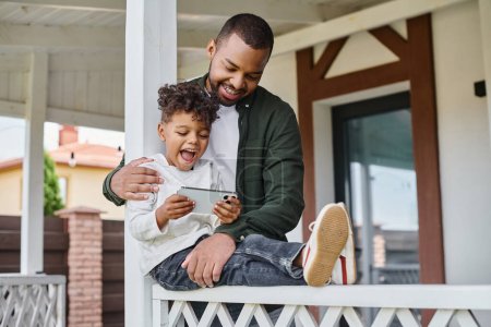 freudig afrikanisch-amerikanischer Vater umarmt aufgeregten Sohn auf Veranda sitzend und Smartphone in der Hand