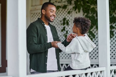 joyful african american father holding hands of excited son sitting on porch on house in suburbs