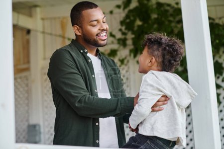 cheerful african american father hugging cute curly son sitting on porch on house in suburbs