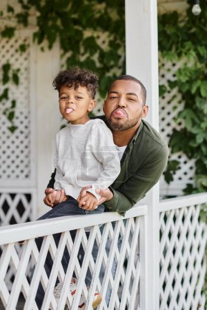 happy african american father and curly son sticking out tongue while sitting near white fence