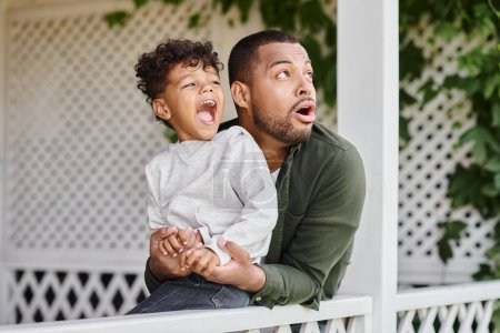 astonished african american father and son looking away surprisingly while sitting near white fence