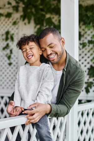 happy african american man in braces hugging curly son while sitting on porch with white fence