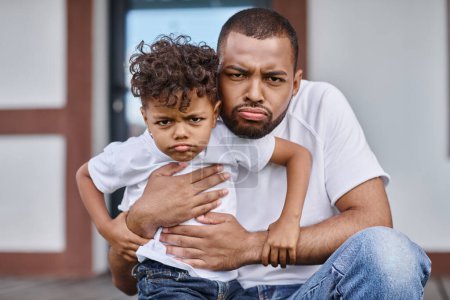 funny portrait of african american son and father looking displeased and sitting on porch of house