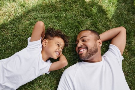 top view of cheerful african american father and cute son lying on green lawn, family photo