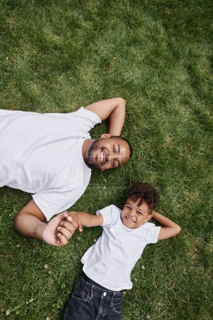 top view of pleased african american father and son lying on green lawn on backyard, family photo