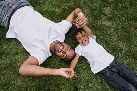 top view of cheerful african american father and son lying on lawn on backyard and holding hands