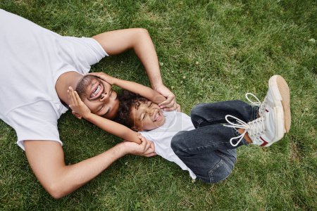 top view of cheerful african american father and son lying on green grass on backyard, family photo