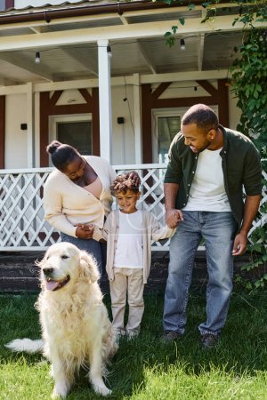 Foto de Momentos felices, padres afroamericanos alegres e hijo sonriendo cerca de perro de la familia en el patio trasero de la casa - Imagen libre de derechos