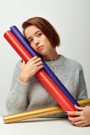 young woman with short hair holding colorful wrapping paper on grey backdrop, Christmas concept