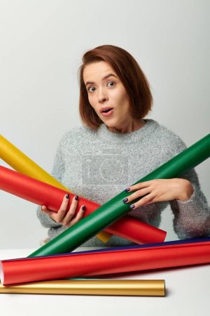 wow emoción, mujer en suéter sosteniendo papel de embalaje de colores en la mesa con la pared gris, Navidad