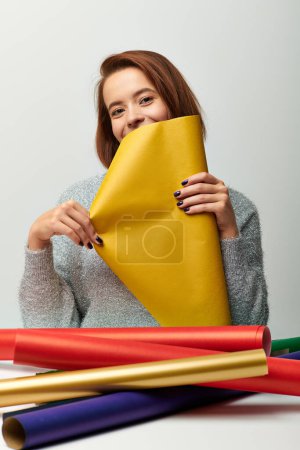 temporada de alegría, mujer joven en suéter sosteniendo papel de regalo de Navidad amarillo sobre fondo gris
