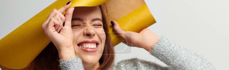 mujer alegre en suéter envolviéndose en papel de regalo amarillo sobre fondo gris, bandera de Navidad